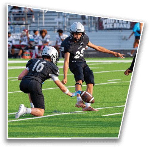 Two people in black football uniforms are holding and kicking a football on a green field while a person in a white and maroon uniform approaches. Description generated by AI