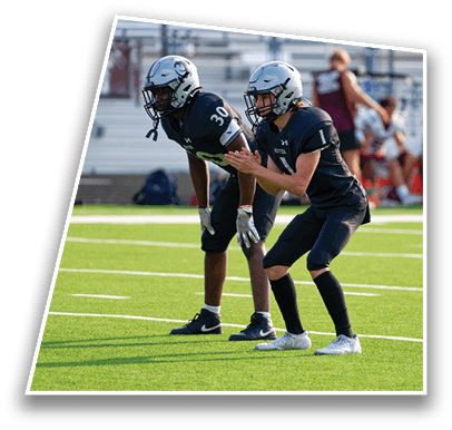 Two people in football uniforms and helmets are positioned on a green field with white yard lines, appearing ready for a play. Description generated by AI