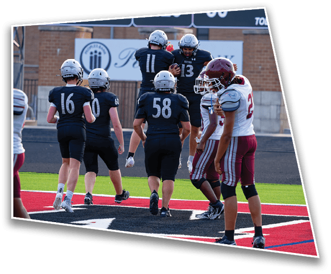 Players in football uniforms are on a field near the end zone with a scoreboard showing a game between Princeton and Denton. Description generated by AI