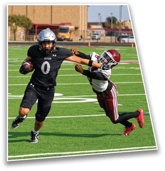 Two people in football uniforms are actively engaged in a play on a green field with white yard lines, with one person attempting to tackle the other who is holding the ball. Description generated by AI