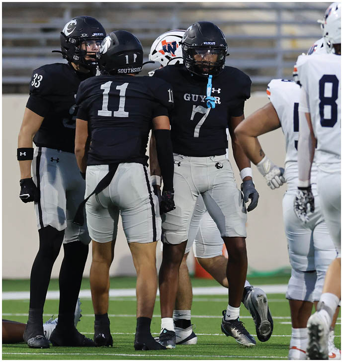 Four football players in black and gray uniforms are standing on a field facing three players in white uniforms during a game. Description generated by AI