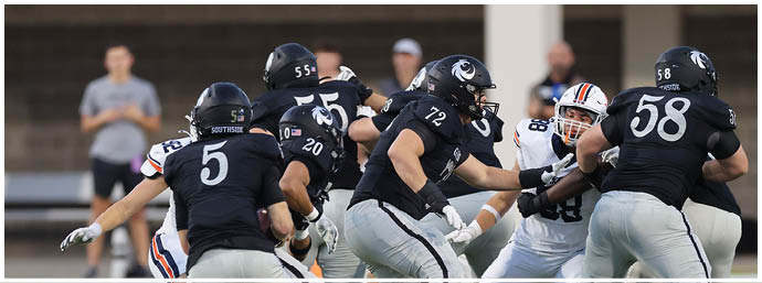 A group of people wearing black football uniforms are engaged in a play on a football field with a scoreboard visible in the background. Description generated by AI