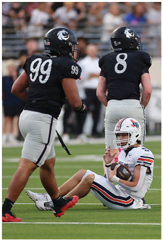 Two football players in black jerseys with numbers 99 and 8 stand on the field while a player in a white uniform with a helmet sits on the ground holding a football. Description generated by AI