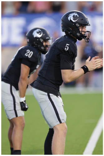 Two people in black football uniforms and helmets are positioned on a field, with one clapping hands and the other crouched behind. Description generated by AI