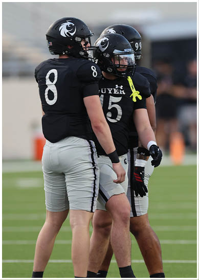Three people wearing black football uniforms and helmets stand together on a green field during a game or practice. Description generated by AI