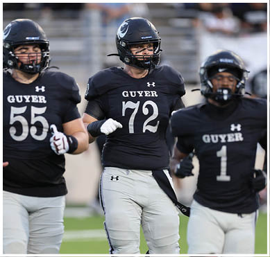 Two people wearing black football uniforms with helmets and gloves are standing on a field, one with the number 72 and the other with the number 55 on their jerseys. Description generated by AI