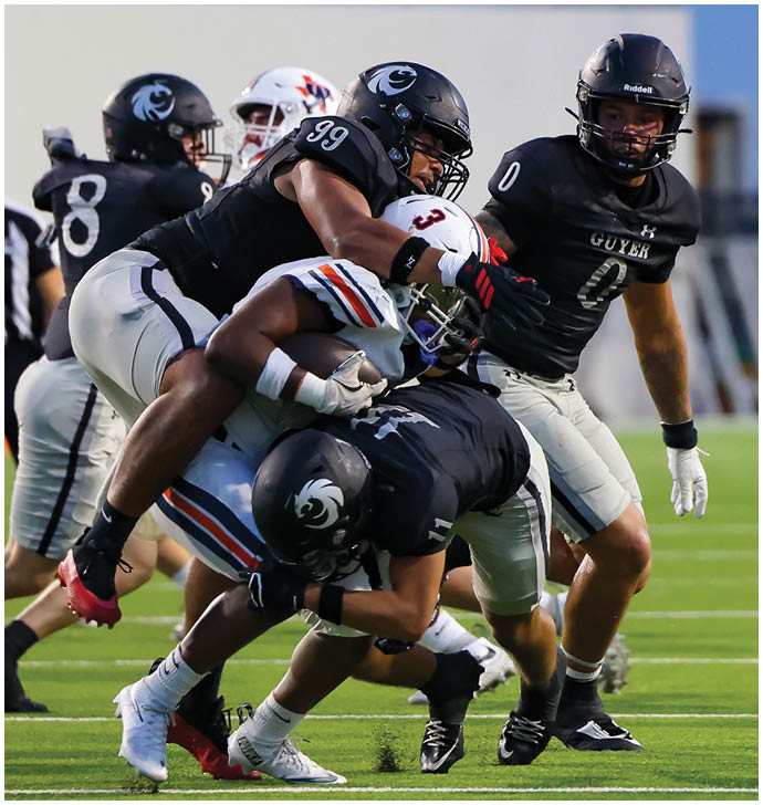 A group of football players in black and white uniforms are tackling a player in a white uniform holding the ball on a green field during a game. Description generated by AI