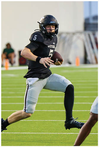A person wearing a black football uniform with the number five is running on a green field while holding a football. Description generated by AI