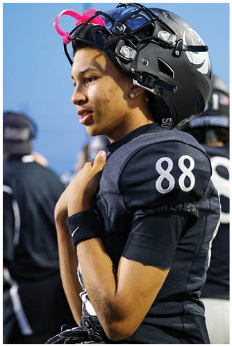 A person wearing a helmet with a pink strap is looking to the right against a blurred background with another person wearing a headset. Description generated by AI