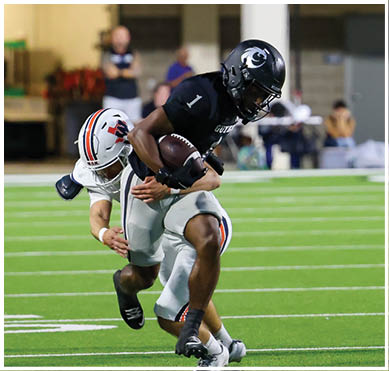 Two football players in black and gray uniforms with helmets are on a green field with white yard lines, one holding a football and the other running nearby. Description generated by AI