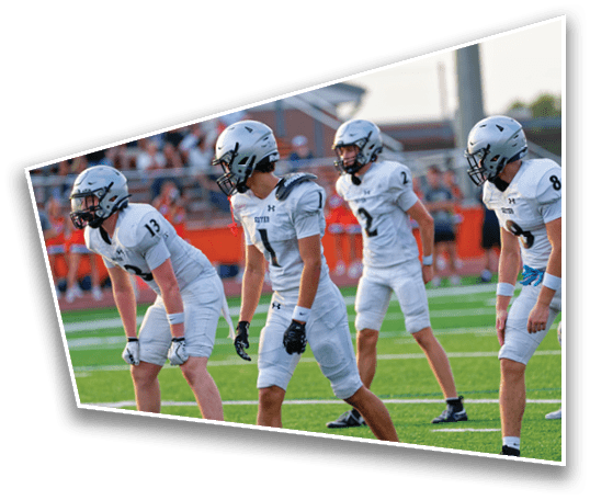 Four people in white football uniforms and helmets are positioned on a green field, appearing ready for a play during a game. Description generated by AI