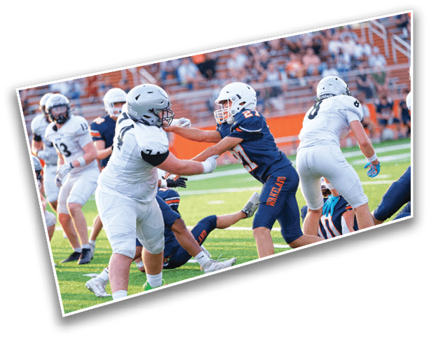 Two teams of people in football uniforms are actively engaged in a game on a green field with spectators in the background. Description generated by AI