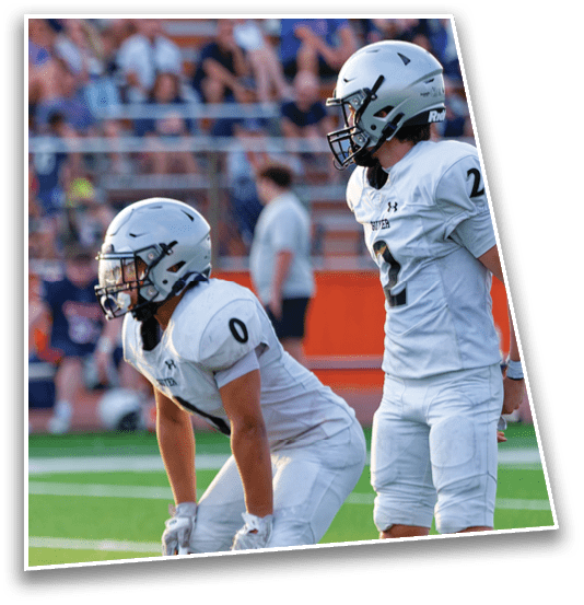Two people in white football uniforms and helmets are on a field with one standing and the other bent forward in a ready position. Description generated by AI