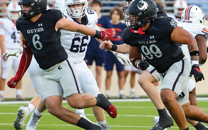Two people in black football uniforms are blocking or engaging with a person in a white football uniform during a game with spectators in the background. Description generated by AI