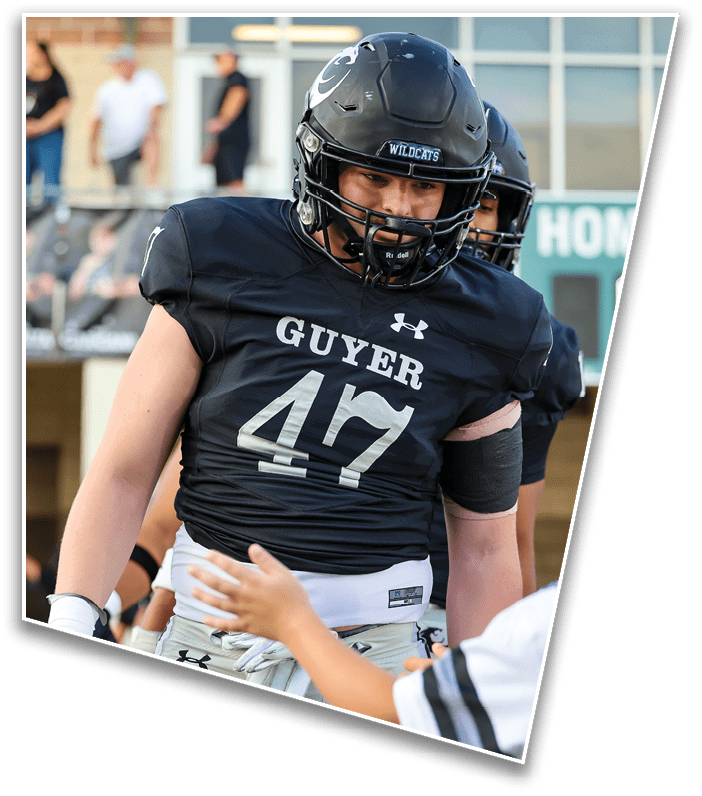A person wearing a black football helmet and black jersey with the word “GUYER" and "WILDCATS" on the helmet is on a sports field with blurred people and a building in the background. Description generated by AI