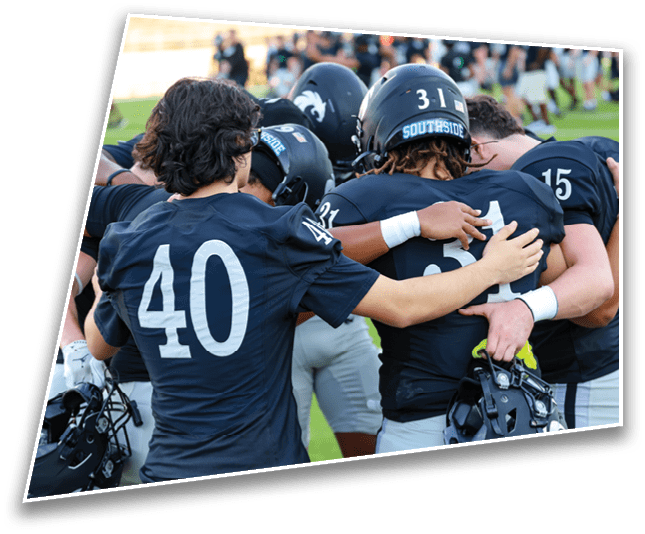 A group of people wearing dark sports uniforms and helmets are huddled together on a grassy field, showing camaraderie and support. Description generated by AI
