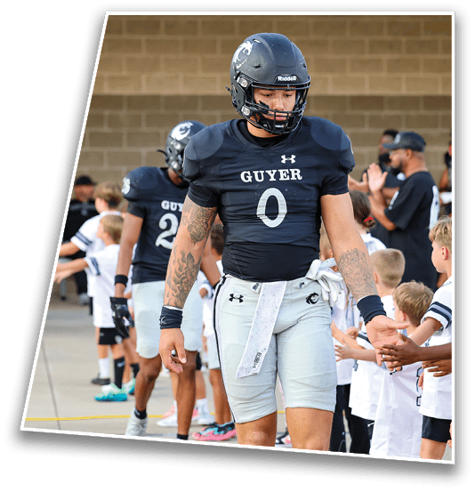 Two people in football uniforms with helmets walk past a line of children reaching out to them, set against a brick wall background. Description generated by AI
