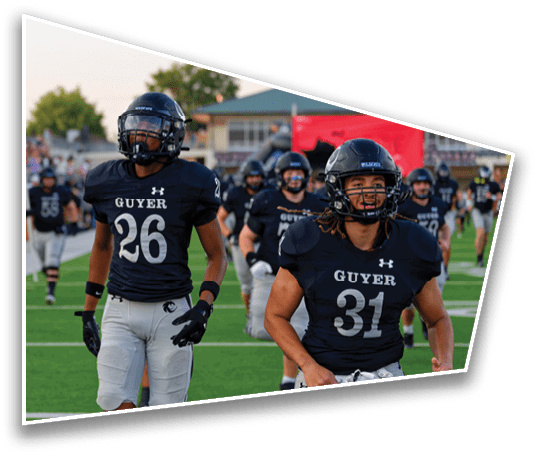 Two people in football uniforms with helmets and gloves are walking on a field with other players and a crowd in the background. Description generated by AI