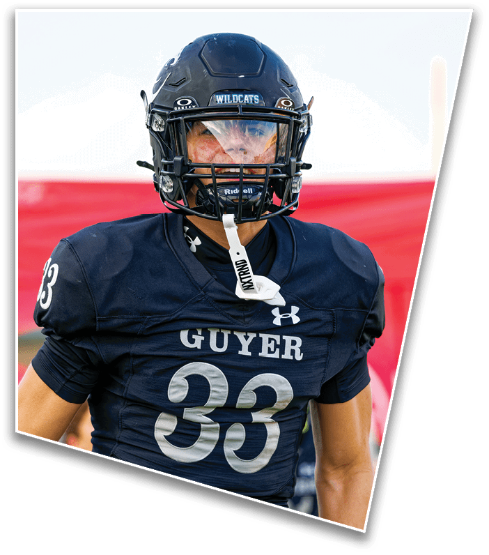 A person wearing a dark blue football uniform with the number 33 and the word “GUYER" on the front is standing on a field with a red banner and other people in the background. Description generated by AI