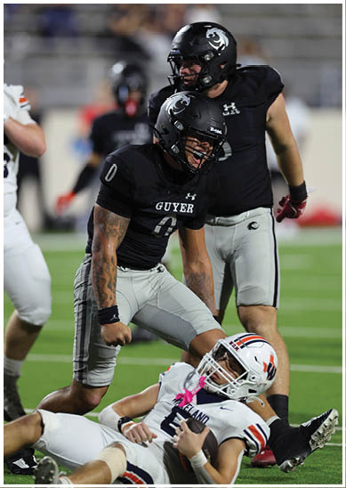 Two people in black football uniforms stand over a person in a white football uniform lying on the field during a game. Description generated by AI
