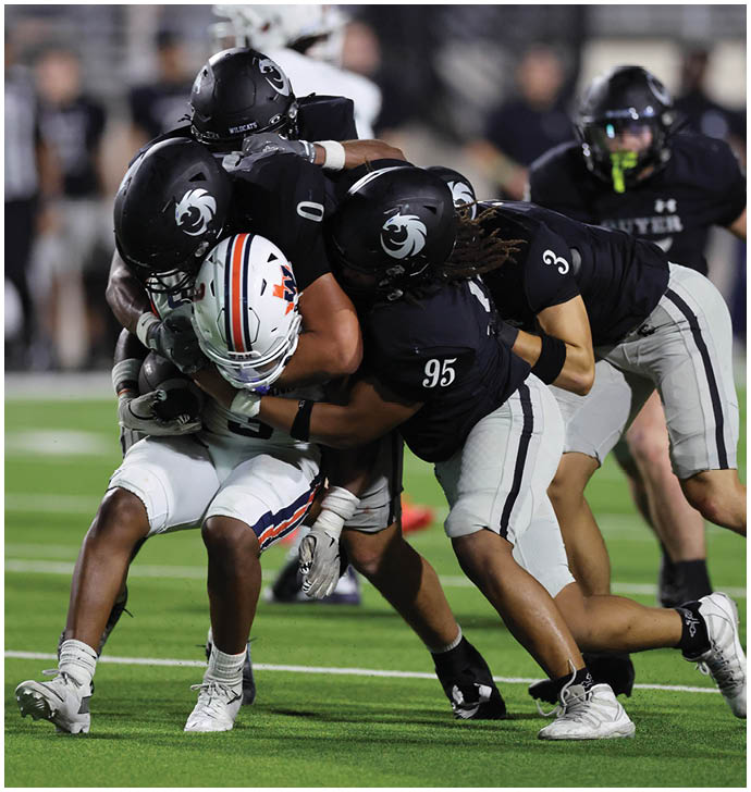 A group of football players in black uniforms tackle a player in a white uniform holding the ball on a green field during a game. Description generated by AI
