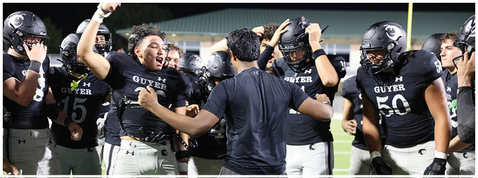 A group of people wearing sports shorts and cleats stand closely together on a grassy field, some holding helmets. Description generated by AI