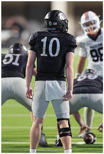 An adult wearing a black football jersey with the number 10 and a knee brace stands on a football field with other players in the background. Description generated by AI