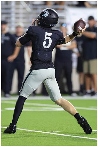 A person wearing a black football uniform with the number 5 is preparing to throw a football on a green field with blurred spectators in the background. Description generated by AI