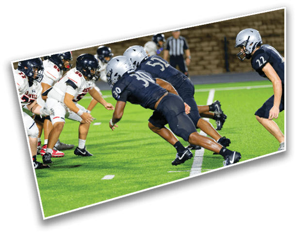 Two opposing American football teams in helmets and uniforms are lined up facing each other on a green field with a referee in the background. Description generated by AI