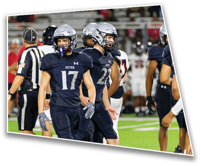 Several football players in dark uniforms and helmets stand on a field during a game with a referee and blurred spectators in the background. Description generated by AI