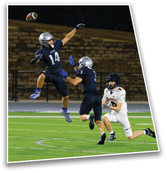 Two people in dark sports uniforms are actively reaching for a football in midair while a person in a white sports uniform runs nearby on a football field. Description generated by AI