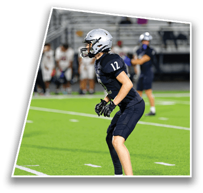 A person wearing a dark football uniform and silver helmet is standing in a ready position on a green football field with other players and spectators in the background. Description generated by AI