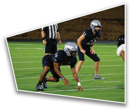 Two people in football uniforms and helmets are positioned on a field with one person in a referee uniform standing behind them. Description generated by AI