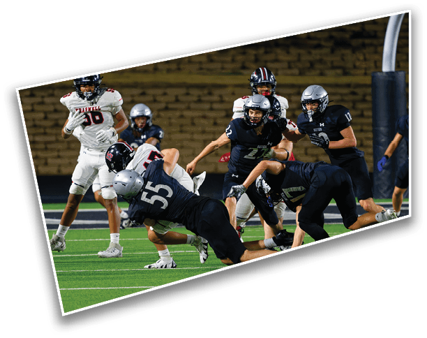 Several people wearing football helmets and uniforms are actively engaged in a play on a green football field with a brick wall background. Description generated by AI