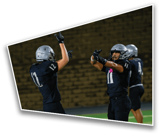 Two people in football uniforms and helmets are celebrating on a field with one raising both hands and the other pointing forward. Description generated by AI