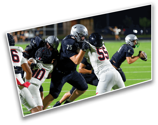 Several people in football uniforms are actively engaged in a play on a well-lit field during a nighttime game. Description generated by AI