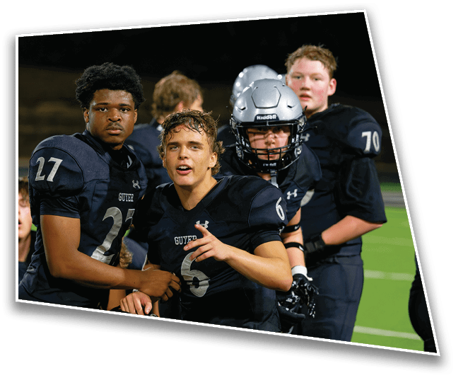 A group of football players in dark uniforms and helmets stand closely together on a field at night, with one player pointing and speaking. Description generated by AI