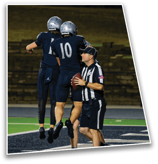 Two people in dark sports uniforms jump in celebration near a referee holding a football while two people in white sports uniforms stand nearby on a football field. Description generated by AI