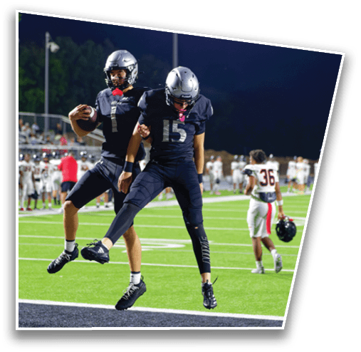 Two people wearing black football uniforms and silver helmets are on a football field at night, with one holding a football and the other standing nearby. Description generated by AI