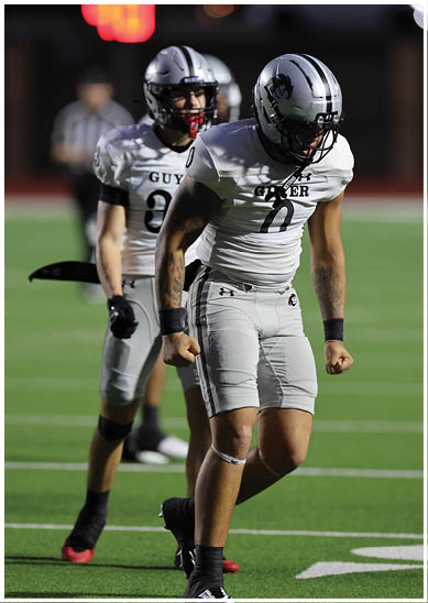 Two people in football uniforms and helmets stand on a field with one person clenching fists and the other standing behind. Description generated by AI