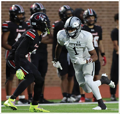 Two football players in contrasting uniforms are engaged in a play on a field with other players and a brick wall in the background. Description generated by AI