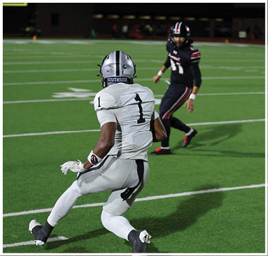 A person wearing a white football uniform with the number one is running on a green field near the sideline while another person in dark clothing is visible in the background. Description generated by AI