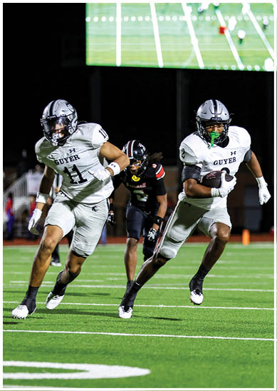 Two people in white football uniforms with “Guyer" on their jerseys are running on a football field at night while a person in a black uniform follows behind them. Description generated by AI