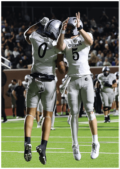 Two football players in white uniforms jump and celebrate on a field with a crowd and other players in the background. Description generated by AI