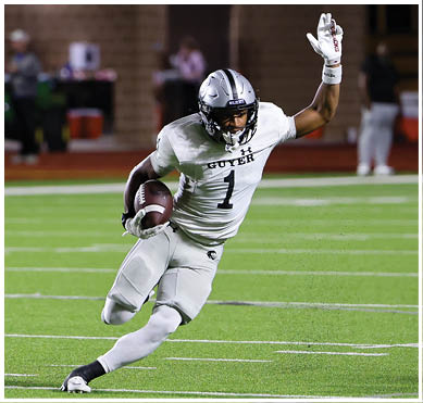 A person wearing a silver helmet and white football uniform with the number one is running on a football field while holding a football and raising one arm. Description generated by AI
