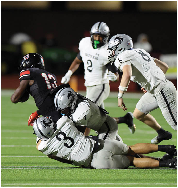 Two people in gray football uniforms tackle a person in a black uniform on a green field during a game. Description generated by AI