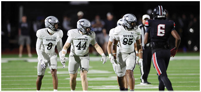 Four people in white football uniforms are walking on a field toward a person in a black football uniform during a nighttime game. Description generated by AI