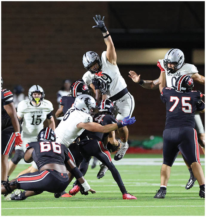 A group of people in football uniforms are actively engaged in a play on a football field with one person jumping and others attempting to block or tackle. Description generated by AI