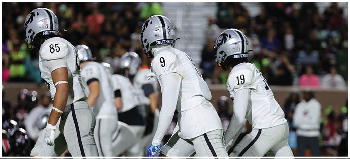 Three football players in white uniforms and helmets stand on a field with a blurred crowd in the background. Description generated by AI