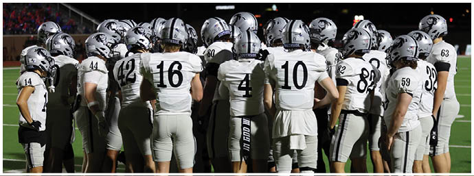 A group of people wearing white football uniforms and helmets are huddled together on a football field at night. Description generated by AI